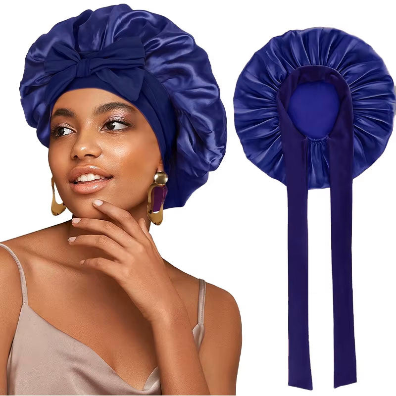 A woman wearing a navy blue satin sleeping bonnet with a tie up at the headband, smiling and posing.